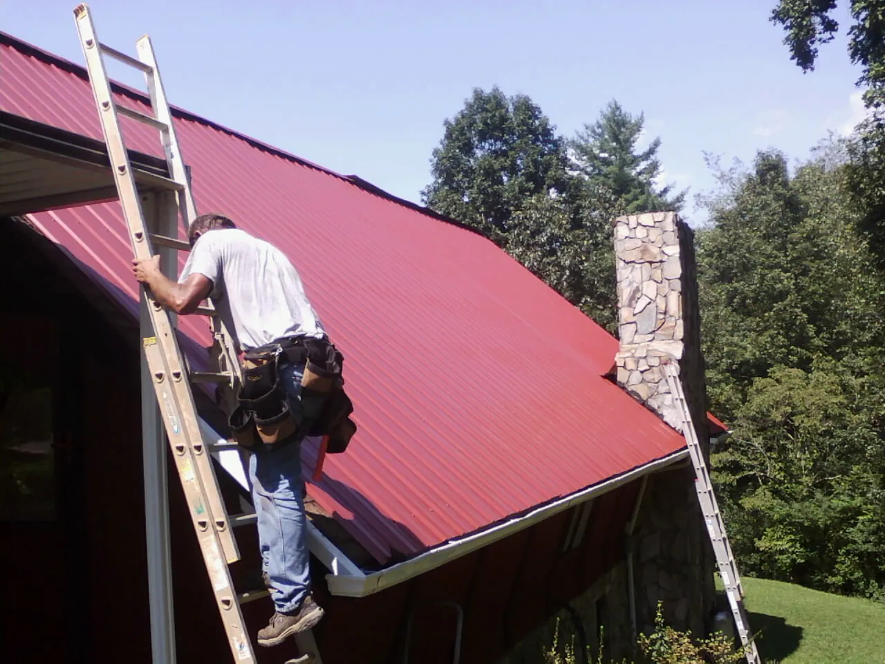 Roofer on a ladder beside a red metal roof and stone chimney — Soffit & Fascia Repair by Roofing Brothers in Wood Ridge
